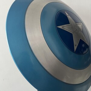 May include: A blue and silver shield with a central star design. The shield has a circular shape with concentric rings of blue and silver. The star is silver and has a five-pointed design. The shield is a prop.