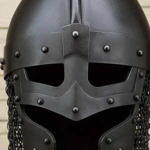 May include: A close-up of a black Viking helmet. The helmet features a riveted face guard with eye openings and a chainmail neck guard. The helmet is made of metal and has a matte finish.