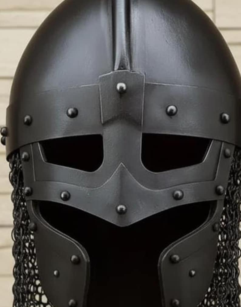 May include: A close-up of a black Viking helmet. The helmet features a riveted face guard with eye openings and a chainmail neck guard. The helmet is made of metal and has a matte finish.