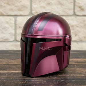 Pu&ograve; includere: Un casco Mandaloriano bordeaux con visiera e dettagli neri. Il casco ha una finitura metallica e le lettere "M.F.C." sono visibili sul lato. Il casco &egrave; esposto su una superficie di legno.