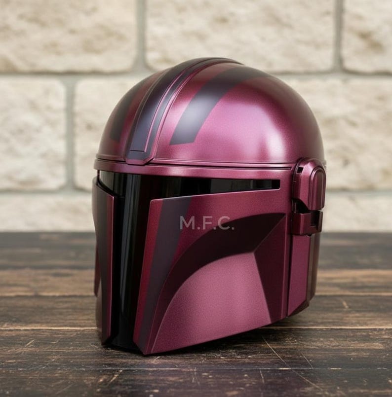 Pu&ograve; includere: Un casco Mandaloriano bordeaux con visiera e dettagli neri. Il casco ha una finitura metallica e le lettere "M.F.C." sono visibili sul lato. Il casco &egrave; esposto su una superficie di legno.