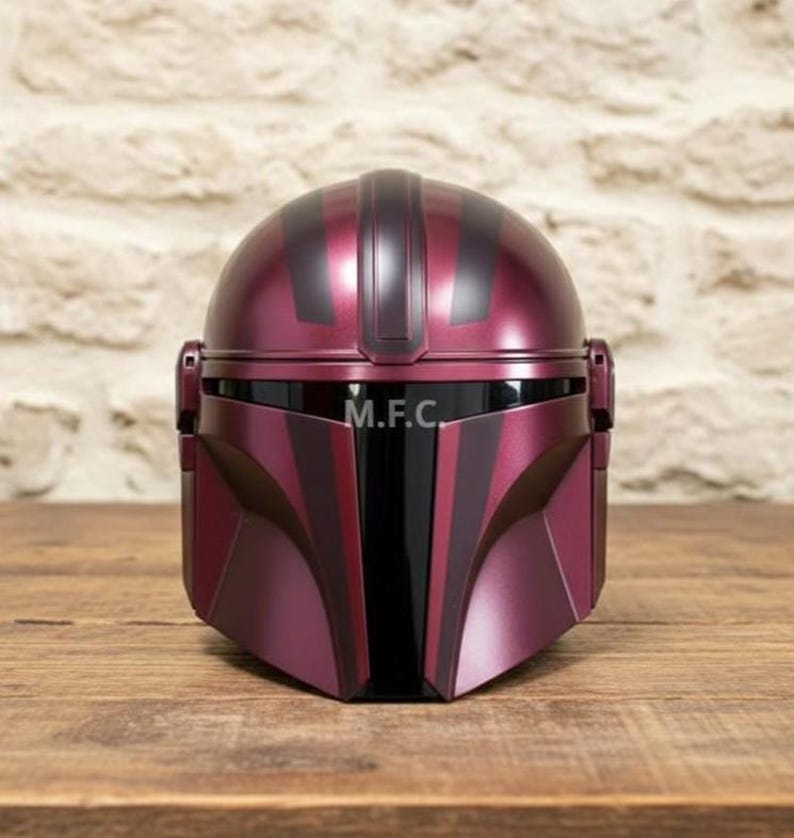 Pu&ograve; includere: Un casco Mandaloriano bordeaux con dettagli neri e le iniziali "M.F.C." sulla visiera. Il casco &egrave; realizzato in un materiale lucido e poggia su una superficie di legno. Il casco ha una caratteristica visiera a forma di T.