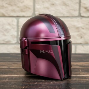 Pu&ograve; includere: Un casco bordeaux con dettagli neri e la scritta "M.F.C." sul lato. Il casco ha una finitura liscia e lucida, con una visiera e un design arrotondato. &Egrave; appoggiato su una superficie di legno.