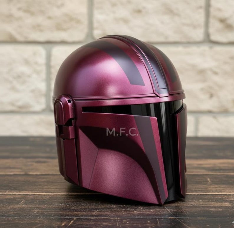 Pu&ograve; includere: Un casco bordeaux con dettagli neri e la scritta "M.F.C." sul lato. Il casco ha una finitura liscia e lucida, con una visiera e un design arrotondato. &Egrave; appoggiato su una superficie di legno.