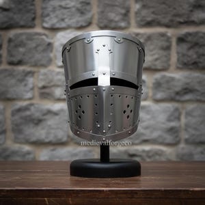 May include: A polished silver medieval helmet with a cross design, rivets, and ventilation holes. The helmet is displayed on a black stand against a stone wall background. The text "medievalforgeco" is visible.