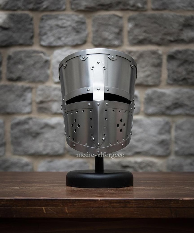 May include: A polished silver medieval helmet with a cross design, rivets, and ventilation holes. The helmet is displayed on a black stand against a stone wall background. The text "medievalforgeco" is visible.