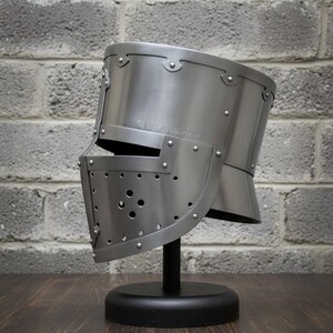 May include: A silver-colored medieval helmet with a visor, riveted construction, and a rounded top. The helmet is displayed on a black stand against a gray brick wall. The text "medievalforgeco" is visible on the side of the helmet.