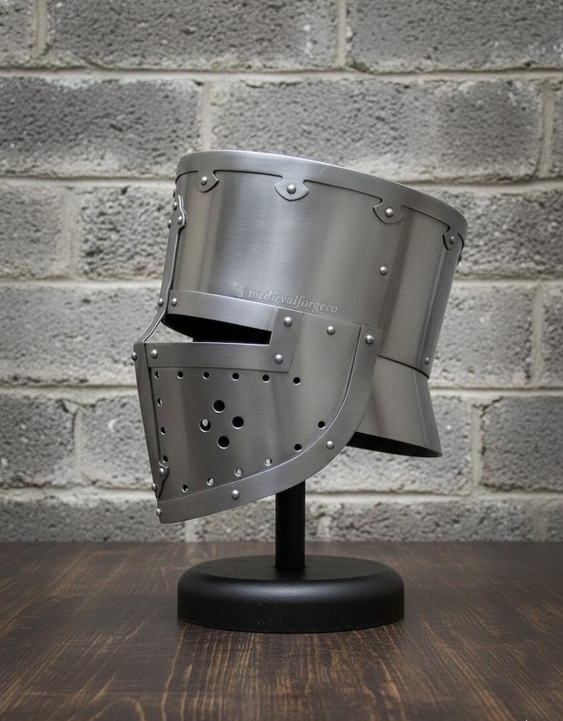May include: A silver-colored medieval helmet with a visor, riveted construction, and a rounded top. The helmet is displayed on a black stand against a gray brick wall. The text "medievalforgeco" is visible on the side of the helmet.