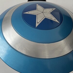 May include: A blue and silver shield with a white star in the center. The shield has a circular design with concentric rings of blue and silver. The star is outlined in black.