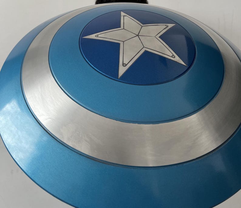 May include: A blue and silver shield with a white star in the center. The shield has a circular design with concentric rings of blue and silver. The star is outlined in black.