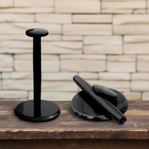 May include: Two black display stands. One stand is upright with a circular base and top. The other stand is a black base with a black cylindrical object and a small black oval object on top. The stands are on a wooden surface.