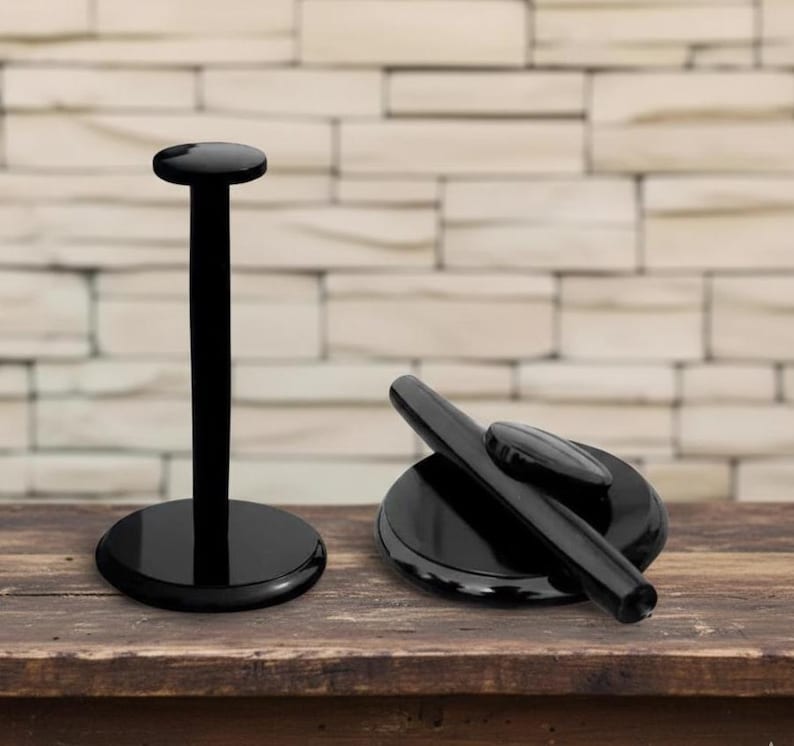 May include: Two black display stands. One stand is upright with a circular base and top. The other stand is a black base with a black cylindrical object and a small black oval object on top. The stands are on a wooden surface.