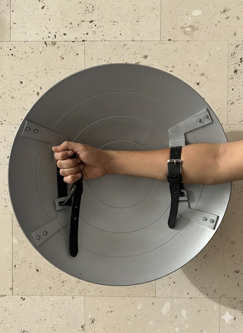 Puede incluir: Un escudo circular plateado con elementos de dise&ntilde;o conc&eacute;ntricos. El escudo tiene un asa y una correa de cuero negro para sujetarlo al brazo. El escudo es un accesorio de utiler&iacute;a o disfraz.