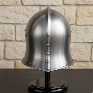 May include: A polished silver medieval helmet with a rounded top and a flared base, riveted with small studs. The helmet is displayed on a black stand against a textured, light-colored brick wall.