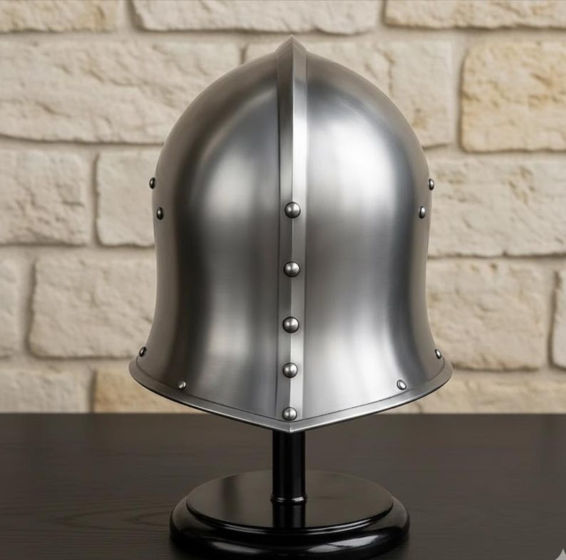 May include: A polished silver medieval helmet with a rounded top and a flared base, riveted with small studs. The helmet is displayed on a black stand against a textured, light-colored brick wall.