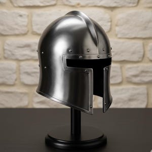 May include: A silver-colored medieval helmet with a visor, rivets, and a central ridge. The helmet is displayed on a black stand with a round base, set against a brick wall background.