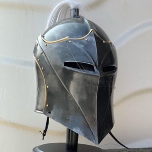 May include: A dark grey medieval helmet with gold detailing and a white plume. The helmet features a pointed visor and is displayed on a black stand. The text "MEDIEVALFORGE.CO" is visible on the side.