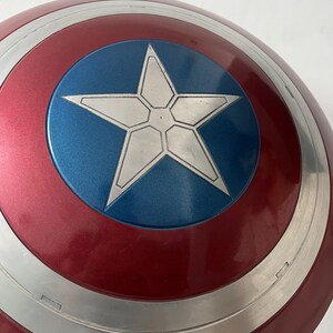 Puede incluir: Un escudo del Capit&aacute;n Am&eacute;rica con una combinaci&oacute;n de colores rojo, blanco y azul. El escudo presenta un c&iacute;rculo azul central con una estrella blanca, rodeado de bandas conc&eacute;ntricas plateadas y rojas. Acabado brillante.