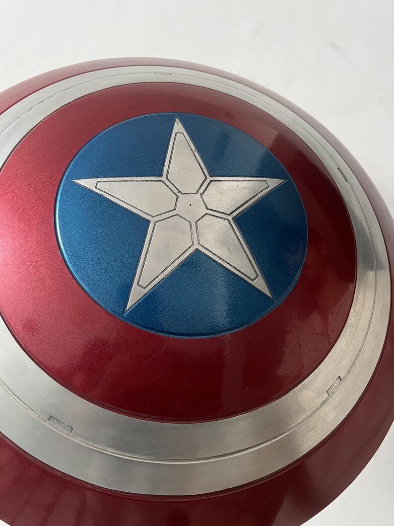 Puede incluir: Un escudo del Capit&aacute;n Am&eacute;rica con una combinaci&oacute;n de colores rojo, blanco y azul. El escudo presenta un c&iacute;rculo azul central con una estrella blanca, rodeado de bandas conc&eacute;ntricas plateadas y rojas. Acabado brillante.