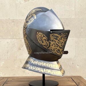 Peut inclure: Casque de style médiéval, gris foncé avec des motifs décoratifs dorés. Le casque est présenté sur un support noir, mettant en valeur son design complexe et son esthétique historique. Le casque est un exemple frappant d'armure historique.