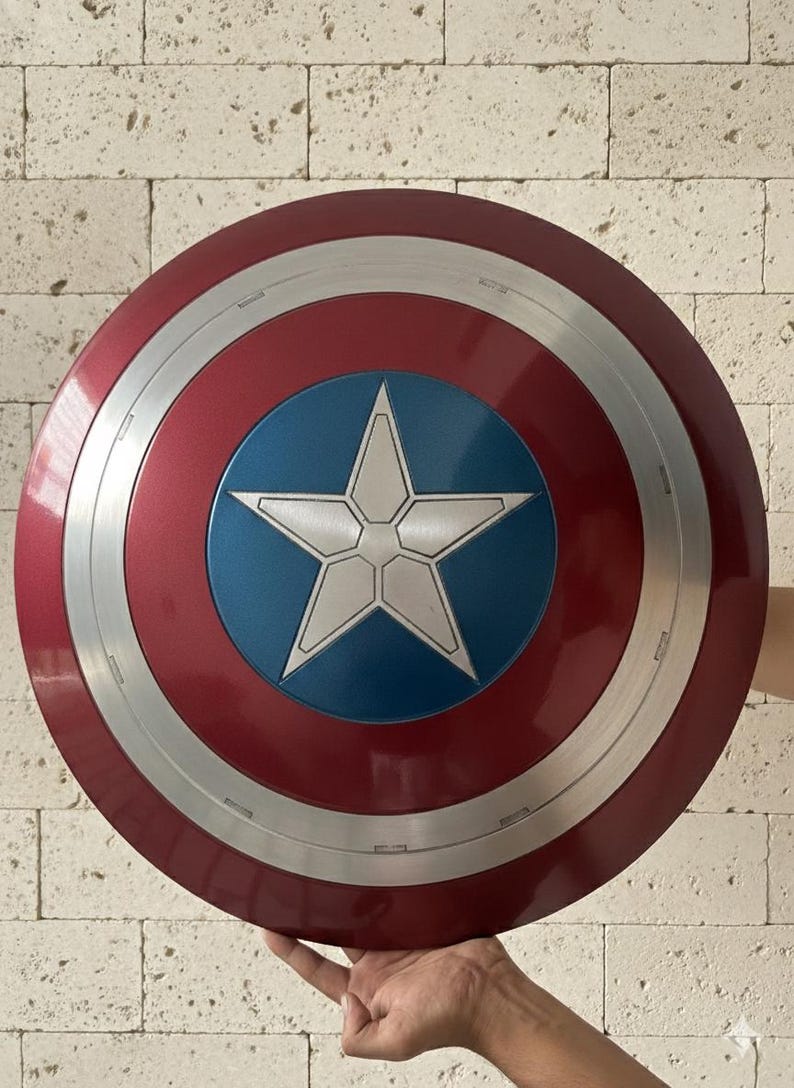 Puede incluir: Un escudo del Capit&aacute;n Am&eacute;rica rojo, blanco y azul. El escudo tiene un dise&ntilde;o circular con un anillo exterior plateado, un &aacute;rea central roja y un centro azul con una estrella blanca. El escudo se sostiene frente a una pared de ladrillos.
