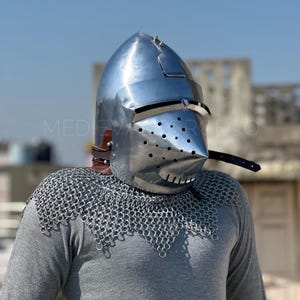 May include: A polished silver medieval helmet with a pointed top and a visor with small holes. The helmet is worn over a chainmail coif and a gray long-sleeve shirt. The text "MEDIEVAL FORGE CO" is visible.