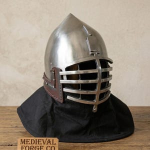May include: A polished metal helmet with a visor featuring horizontal bars. The helmet has a pointed top and is set on a black fabric. A brown leather strap is visible on the side. The wooden sign at the bottom reads "MEDIEVAL FORGE CO."
