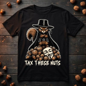 May include: A black t-shirt featuring a graphic of a squirrel dressed in a black hat and cape, standing atop a pile of walnuts and acorns. A white Guy Fawkes mask is nestled among the nuts. The text TAX THESE NUTS is printed in white below.