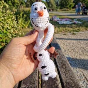 Puede incluir: Mu&ntilde;eco de Olaf de ganchillo hecho a mano. El mu&ntilde;eco de nieve blanco tiene una nariz de zanahoria, ojos negros y pelo de ramitas marrones. El mu&ntilde;eco se sostiene frente a un fondo de parque.