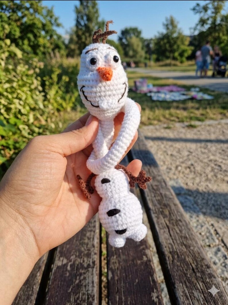 Puede incluir: Mu&ntilde;eco de Olaf de ganchillo hecho a mano. El mu&ntilde;eco de nieve blanco tiene una nariz de zanahoria, ojos negros y pelo de ramitas marrones. El mu&ntilde;eco se sostiene frente a un fondo de parque.