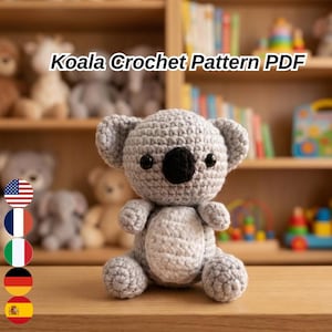 May include: A crocheted koala bear made from grey and white yarn, with black eyes and a black nose. The text "Koala Crochet Pattern PDF" is displayed above the koala. The koala is sitting on a wooden surface.