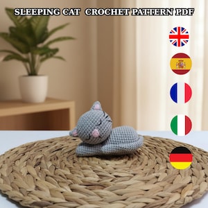 Puede incluir: Un gato durmiente gris tejido a ganchillo con detalles rosas, tumbado sobre una estera tejida. El gato tiene los ojos cerrados y orejas rosas. El texto "SLEEPING CAT CROCHET PATTERN PDF" está en la parte superior.