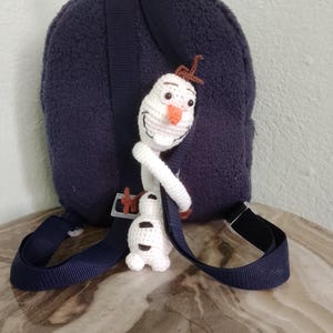 Puede incluir: Una mochila azul marino con correas ajustables y un peque&ntilde;o mu&ntilde;eco de Olaf tejido a ganchillo de color blanco adjunto. El mu&ntilde;eco tiene una nariz de zanahoria y pelo de ramita marr&oacute;n. La mochila tiene una textura difusa.