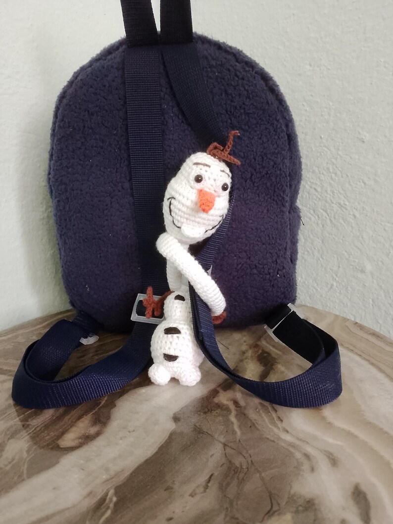 Puede incluir: Una mochila azul marino con correas ajustables y un peque&ntilde;o mu&ntilde;eco de Olaf tejido a ganchillo de color blanco adjunto. El mu&ntilde;eco tiene una nariz de zanahoria y pelo de ramita marr&oacute;n. La mochila tiene una textura difusa.