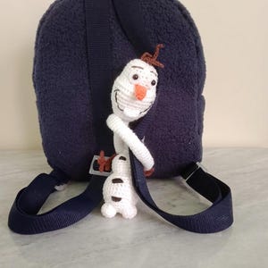 Puede incluir: Una mochila azul marino con una textura de felpa, con un charm de personaje de Olaf tejido a ganchillo. El charm es blanco con una nariz de zanahoria y pelo de ramita marr&oacute;n. La mochila tiene correas azul marino y una hebilla negra.