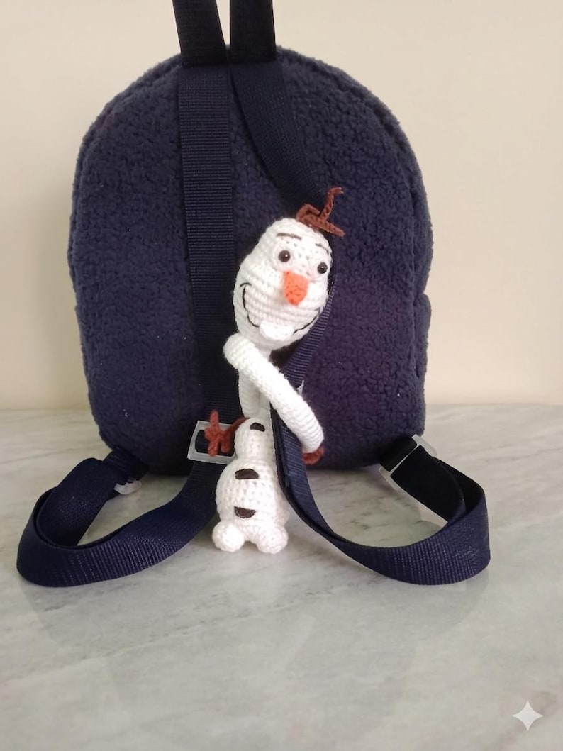 Puede incluir: Una mochila azul marino con una textura de felpa, con un charm de personaje de Olaf tejido a ganchillo. El charm es blanco con una nariz de zanahoria y pelo de ramita marr&oacute;n. La mochila tiene correas azul marino y una hebilla negra.