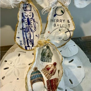 Pode incluir: Enfeites de Natal pintados à mão em conchas de ostra. Um apresenta um quebra-nozes azul e branco, outro diz "Merry & Bright", e o terceiro tem imagens de chapéus de inverno. Cada concha tem uma borda dourada e uma fita branca para pendurar.