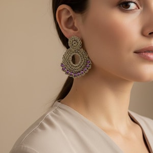 May include: A gold-toned crocheted earring featuring a circular top stud and a larger, open-center circular drop base. The lower edge of the drop is embellished with a row of small, vibrant purple beads, creating a textured and detailed accessory.