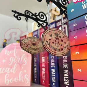 Puede incluir: Carteles colgantes de madera con las palabras "Romance" y "Mamarracheo" delante de una fila de libros. La imagen también incluye un cartel rosa que dice "hot girls read books".