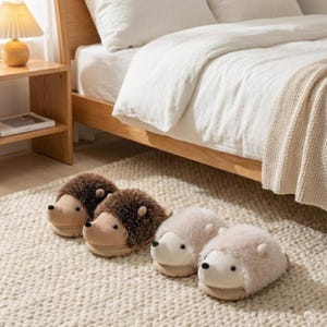以下が含まれることがあります： 寝室に置かれた、ハリネズミの形をした4足のノベルティスリッパ。2足は茶色で顔はタン色、2足は薄いピンク色で顔は白色です。スリッパは、白い寝具のあるベッドの近くにあるクリーム色のラグの上にあります。