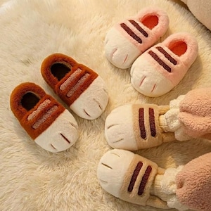 以下が含まれることがあります： 茶色とピンクの色合いの猫の足の形をしたスリッパがいくつかあります。 スリッパは、茶色またはピンクのストライプが付いた白い足のデザインと、柔らかくふわふわした質感を持っています。 スリッパは猫の足のように見えるように設計されています。