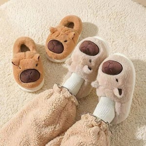 以下が含まれることがあります： 茶色とベージュのクマの形をした居心地の良いスリッパ。濃い茶色の鼻と耳が特徴です。スリッパはクリーム色のラグの上にあります。画像には、お揃いのベージュのパジャマを着た人も写っています。