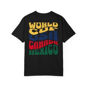 May include: Black t-shirt with a colorful graphic design. The design features the words "WORLD CUP USA CANADA MEXICO" in a retro font, with each word in a different color: yellow, blue, red, and green.