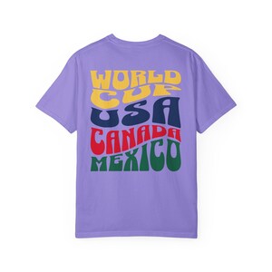 May include: A lilac-colored t-shirt with the words "WORLD CUP USA CANADA MEXICO" in a retro, colorful font. The text is stacked vertically, with each word in a different color: yellow, navy, red, and green. The shirt has a crew neck and short sleeves.
