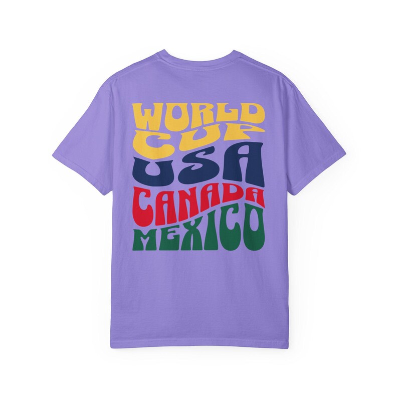 May include: A lilac-colored t-shirt with the words "WORLD CUP USA CANADA MEXICO" in a retro, colorful font. The text is stacked vertically, with each word in a different color: yellow, navy, red, and green. The shirt has a crew neck and short sleeves.
