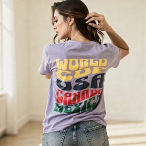 May include: A lilac-colored t-shirt with the words "WORLD CUP USA CANADA MEXICO" in a retro font. The text is in yellow, blue, red, and green. The person is wearing blue jeans.