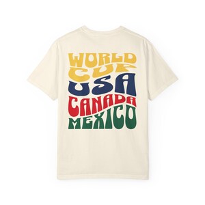 May include: Cream-colored t-shirt with the words "WORLD CUP USA CANADA MEXICO" in a retro, colorful font. The text is stacked and curved, with each word in a different color: yellow, blue, red, and green.