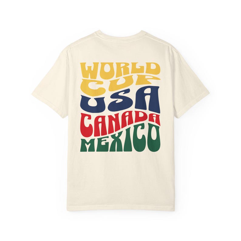 May include: Cream-colored t-shirt with the words "WORLD CUP USA CANADA MEXICO" in a retro, colorful font. The text is stacked and curved, with each word in a different color: yellow, blue, red, and green.