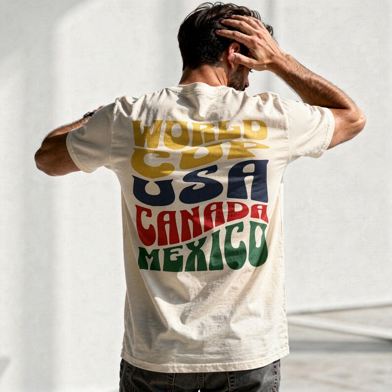 May include: Cream-colored t-shirt with the words "WORLD CDR USA CANADA MEXICO" in a retro font. The text is in yellow, blue, red, and green. The shirt is short-sleeved and has a crew neck.