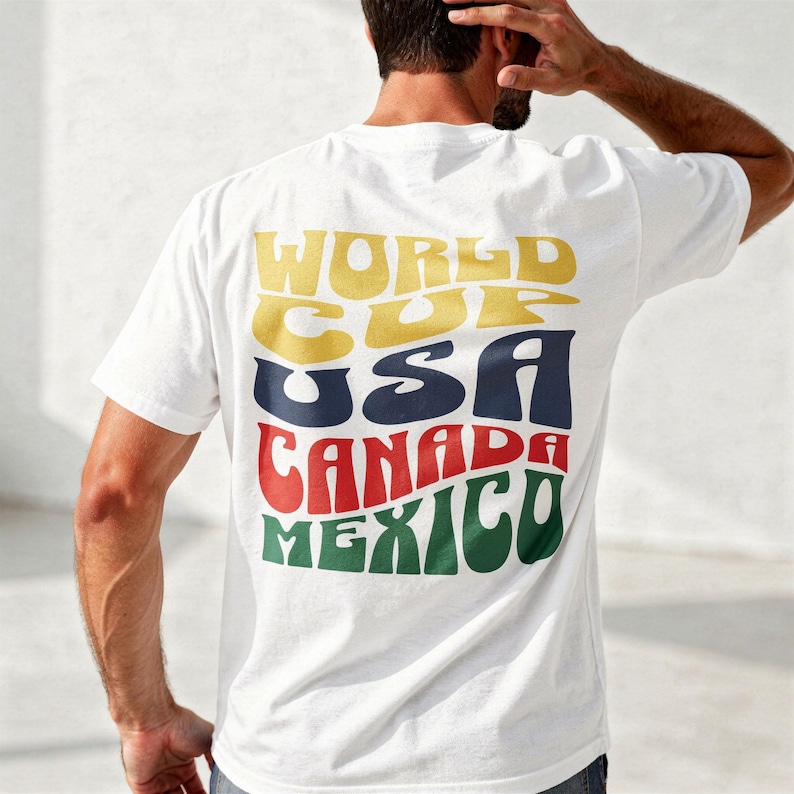 Könnte beinhalten: Weißes T-Shirt mit Retro-Design. Auf der Rückseite des T-Shirts stehen die Worte "WORLD CUP USA CANADA MEXICO" in Gelb, Blau, Rot und Grün. Das T-Shirt hat einen Rundhalsausschnitt und kurze Ärmel.